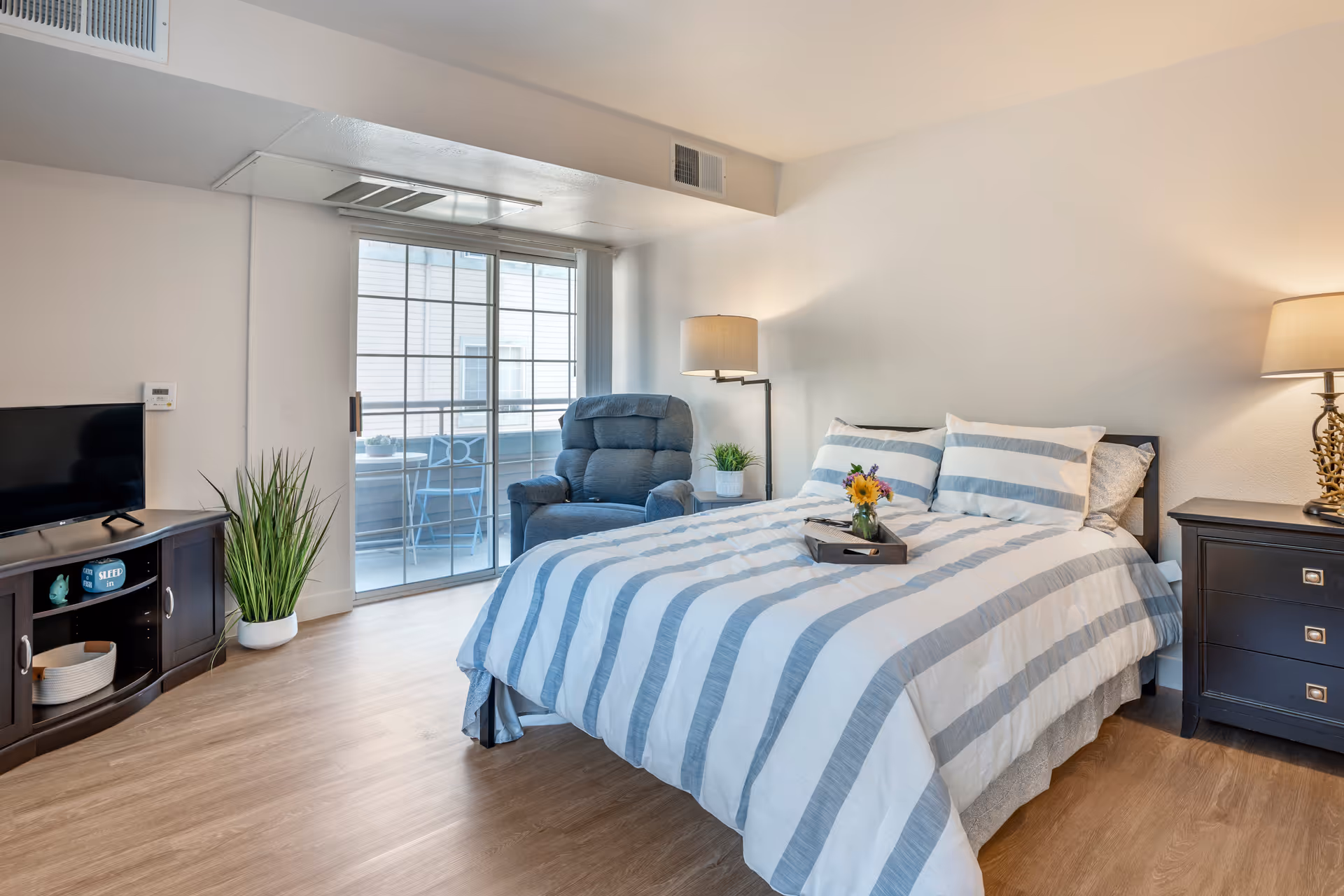 A bright bedroom with a large bed covered in a white and gray striped comforter, two matching pillows, and a tray with a vase of flowers on it. To the right of the bed is a dark wooden nightstand with a lamp. Next to the bed is a blue upholstered armchair and a floor lamp. On the left side of the room is a dark wooden TV stand with a flat-screen TV and a potted plant on the floor. A sliding glass door leads to a small balcony with outdoor furniture. The room has light wood flooring and white walls.