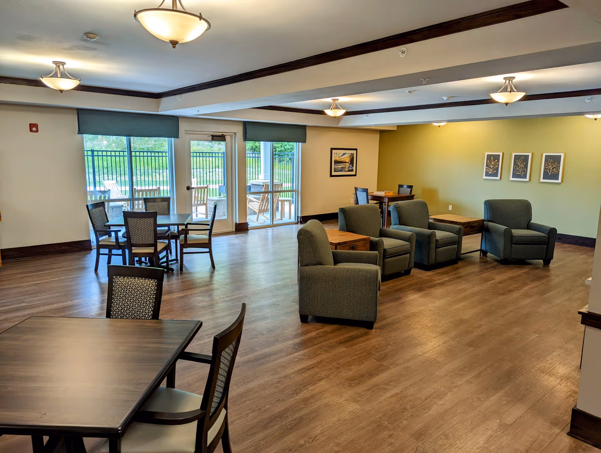 A spacious common area in a senior living facility featuring several tables with chairs and a seating area with armchairs and side tables. Large windows and glass doors provide a view of an outdoor patio with wooden chairs and greenery beyond. The room has wood flooring, light-colored walls with a green accent wall, and ceiling lights.
