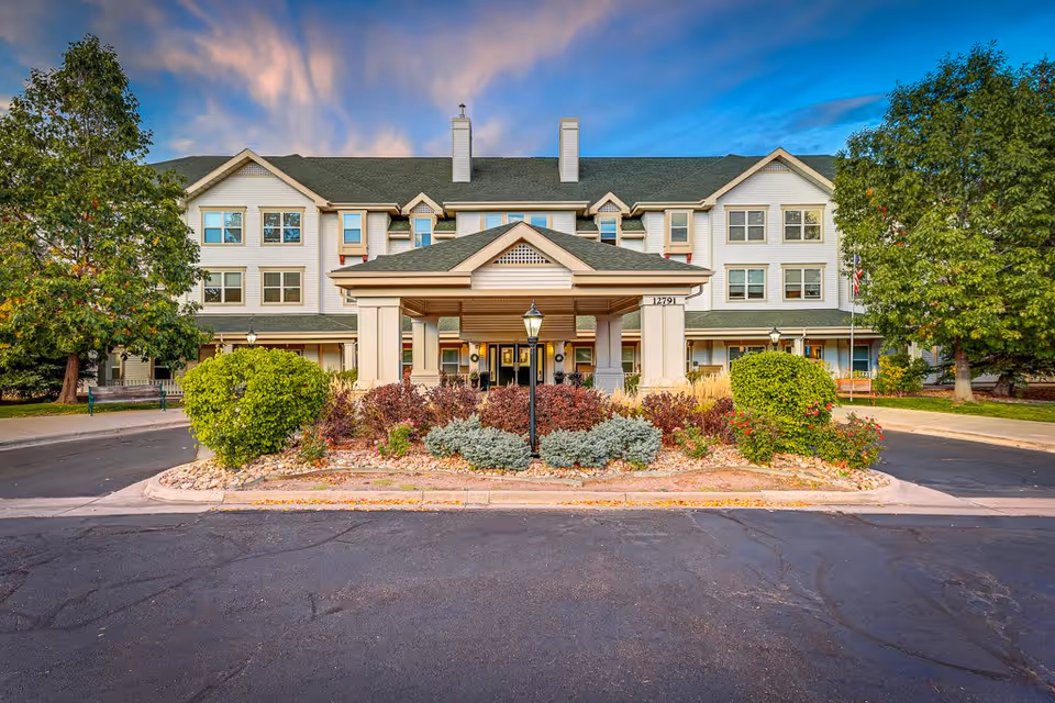 Front exterior view of Woodlake Legacy Senior Living building with a covered entrance, landscaped bushes, trees, and a paved driveway under a partly cloudy sky at sunset.
