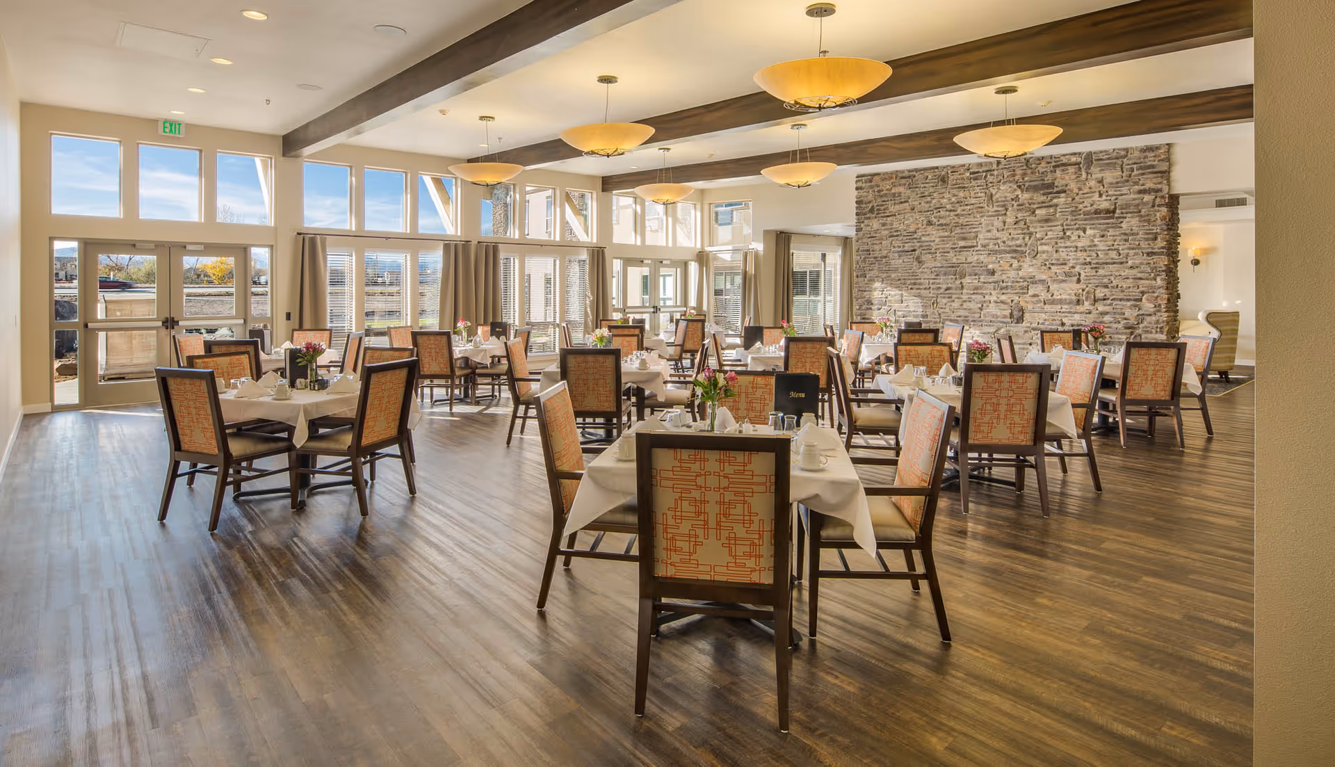 A spacious and well-lit dining room with multiple tables covered in white tablecloths, each set with cups, glasses, and floral centerpieces. The room features large windows allowing natural light to fill the space, wooden beams on the ceiling, modern hanging light fixtures, and a stone accent wall.