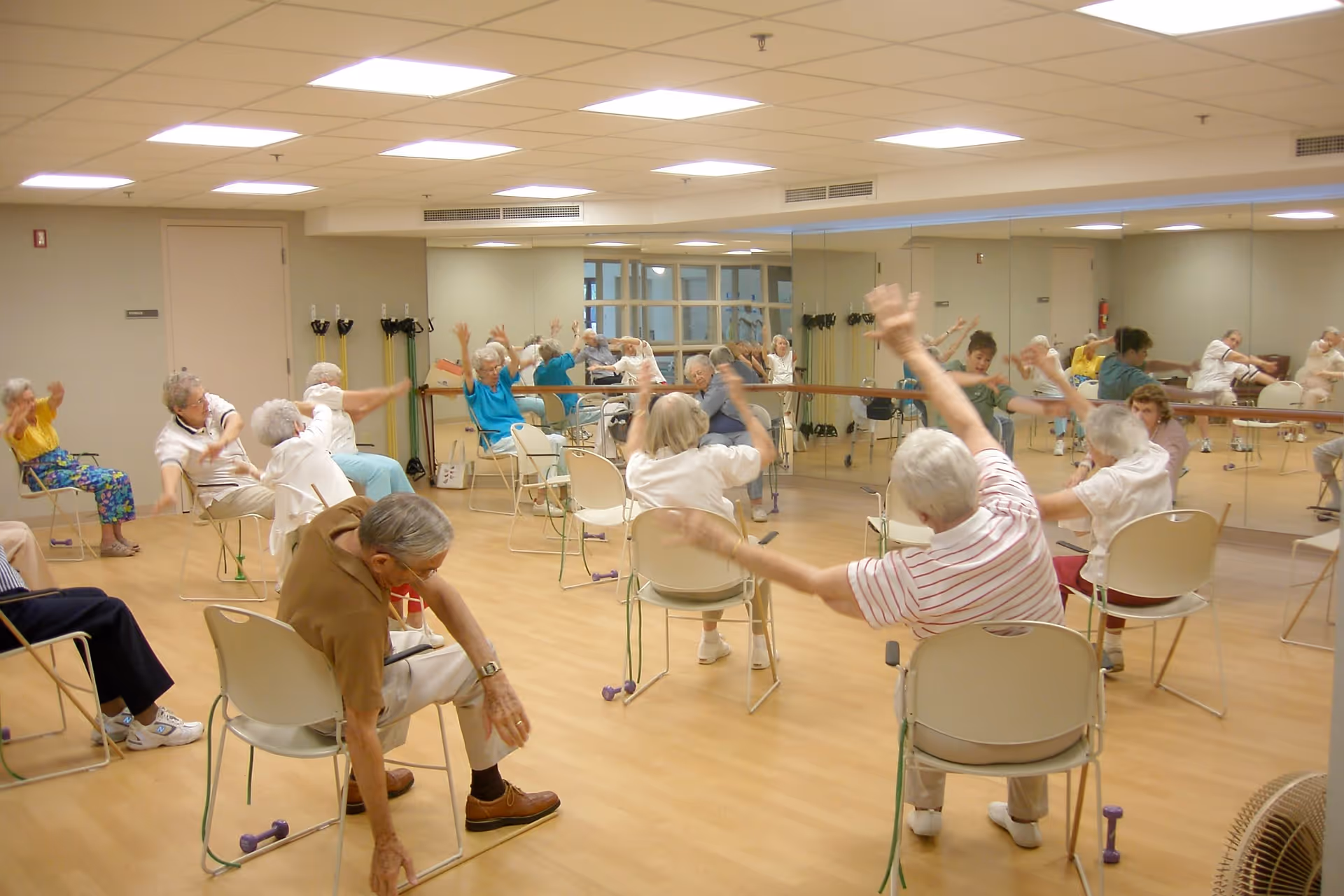 A group of elderly people seated on chairs in a spacious room with wooden floors and mirrored walls, participating in a seated exercise class with arms raised and stretching.