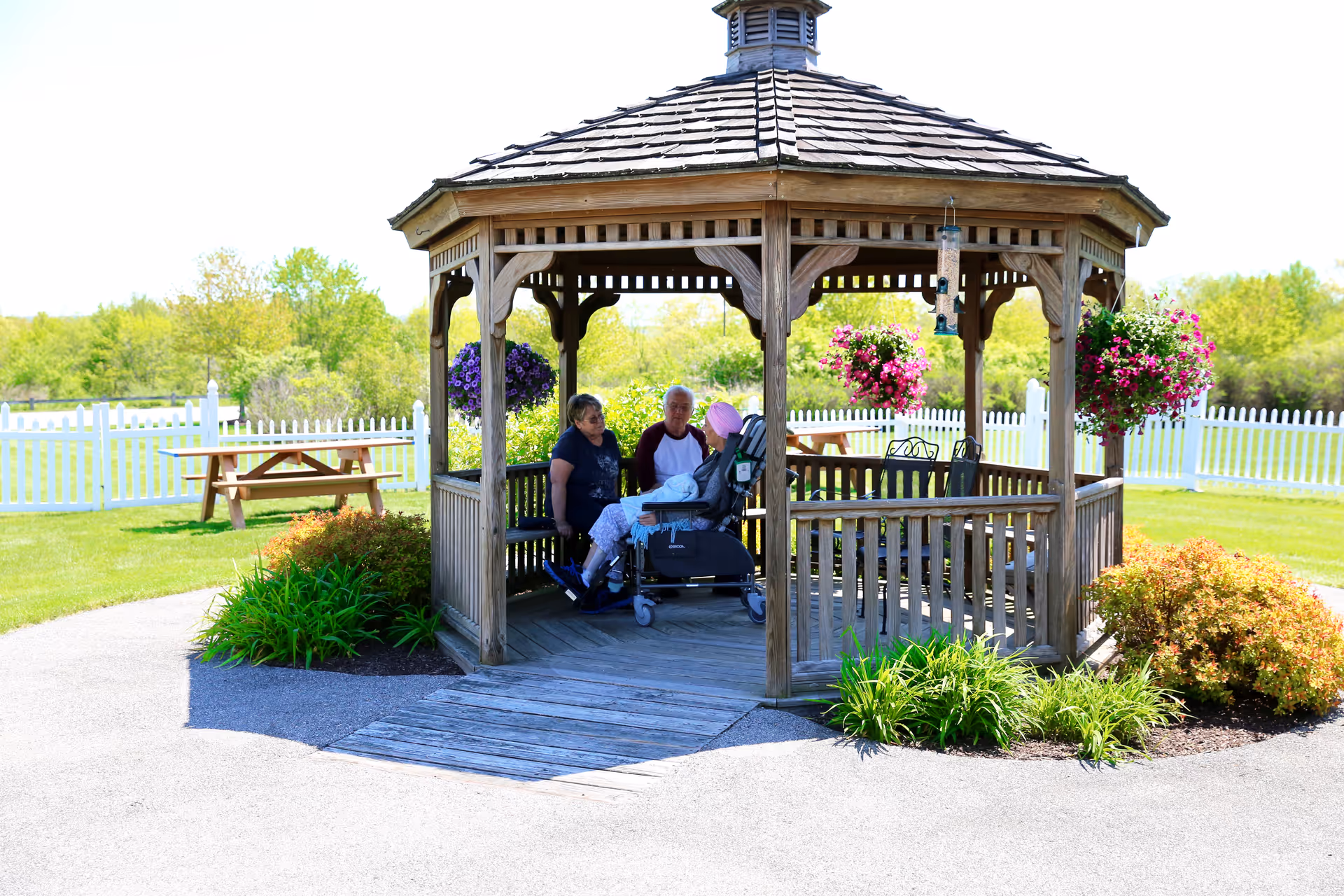 Three elderly people sitting and conversing inside a wooden gazebo in a garden area with green grass, flowering plants, and a white picket fence in the background on a sunny day.