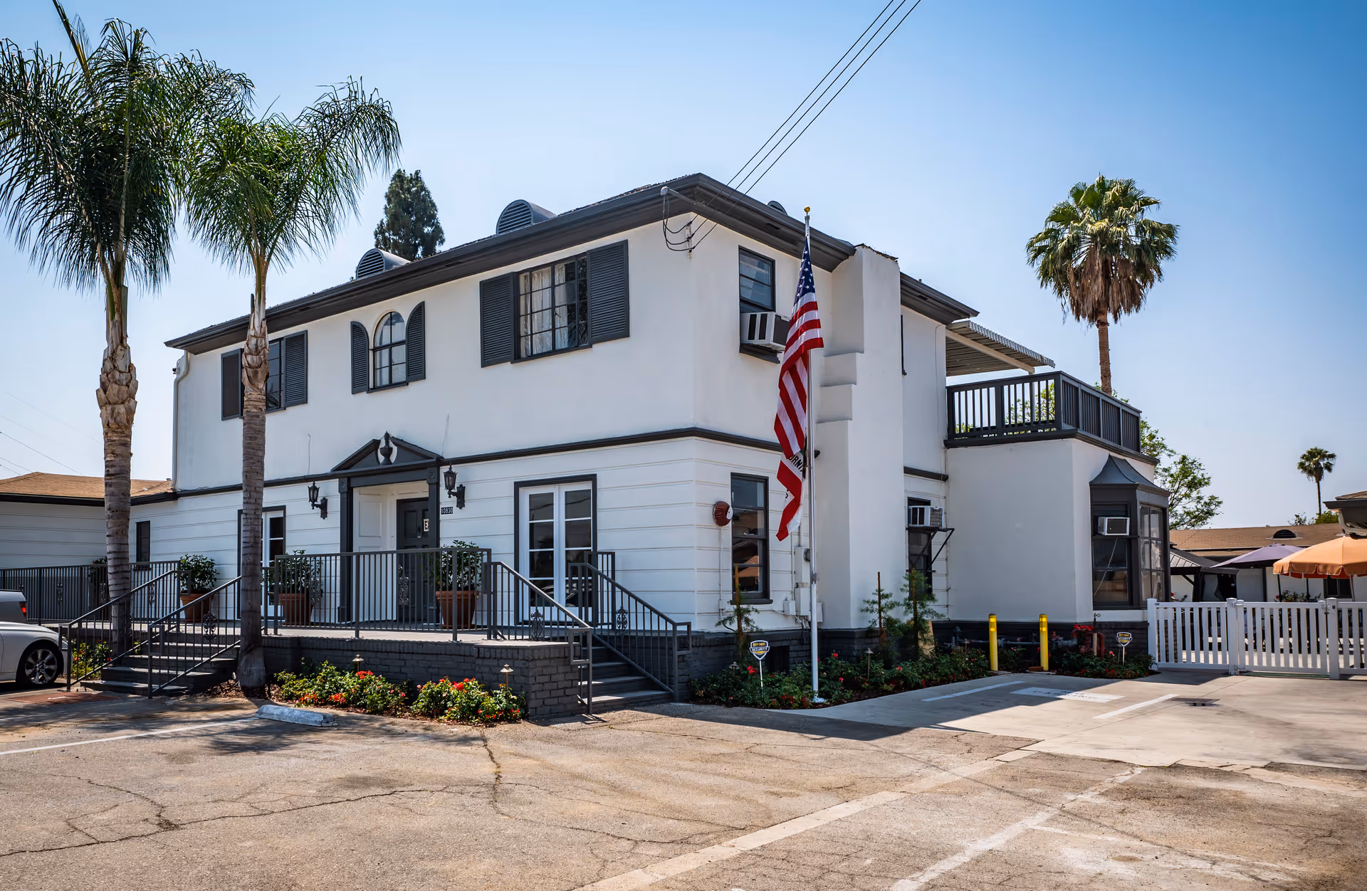 Two-story white building with palm trees, an American flag, and a small parking area in front.