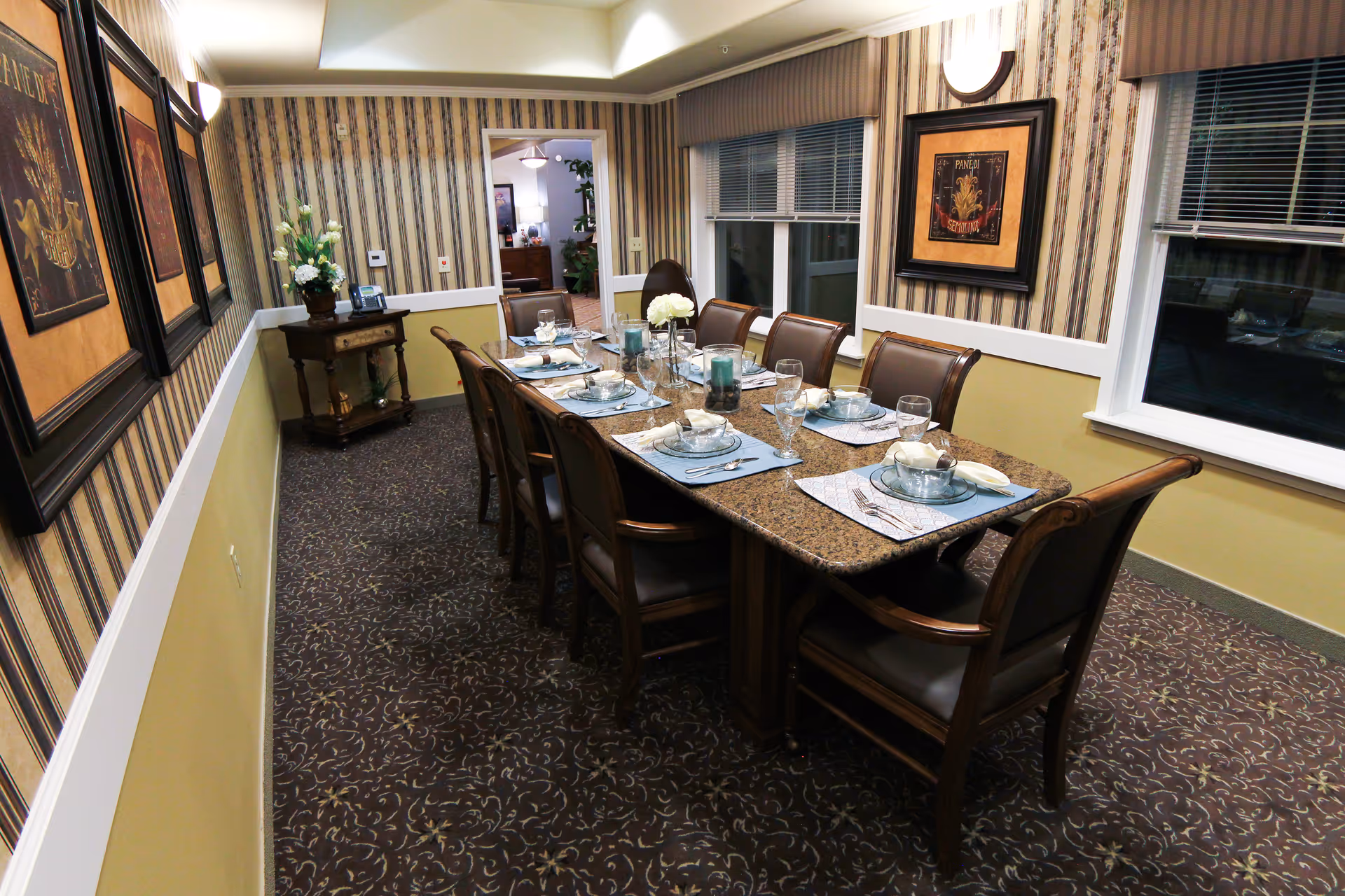 A formal dining room with a long table set for eight, framed artwork, and patterned wallpaper.