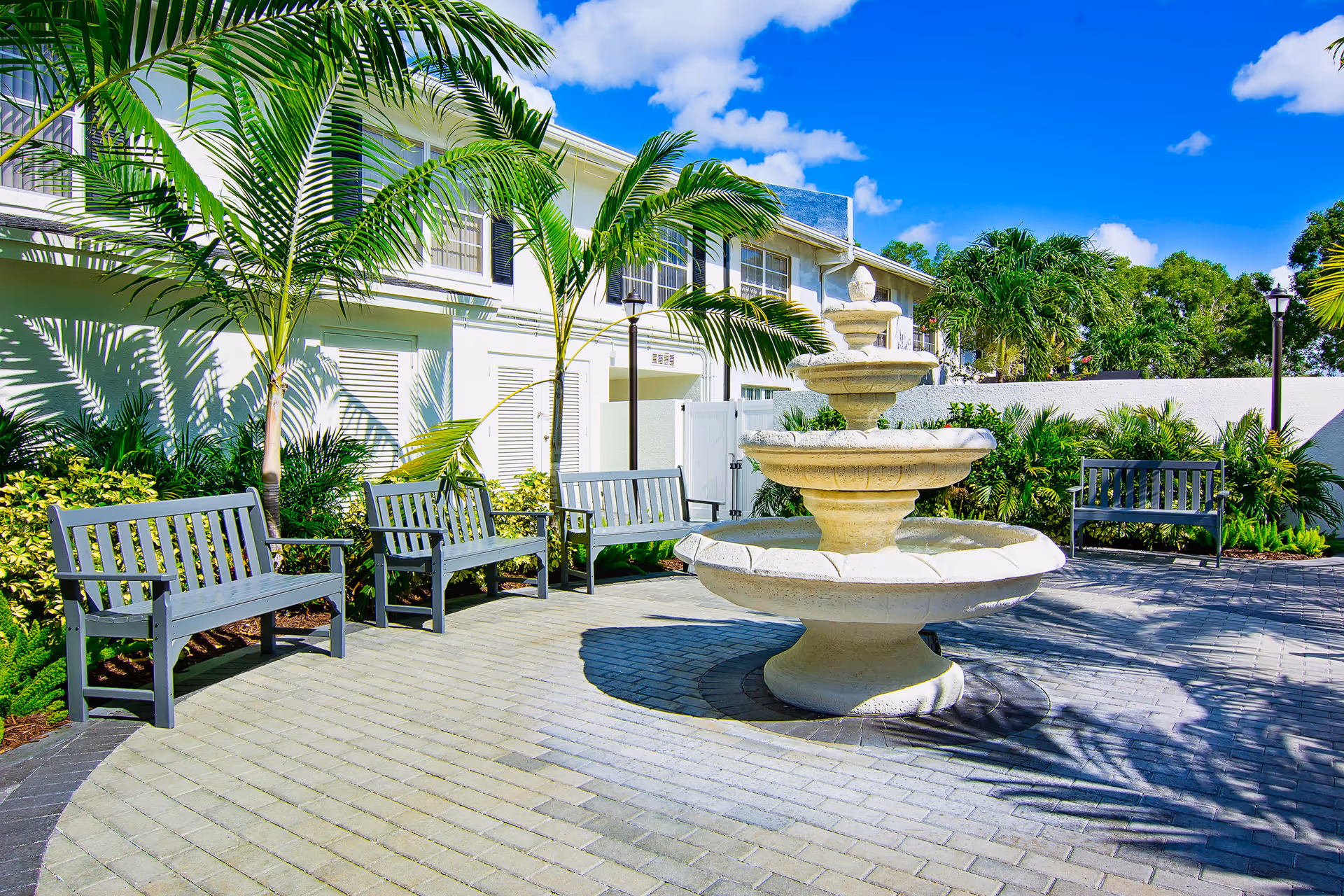 Sunny courtyard with a three-tiered stone fountain surrounded by benches, palm trees, and a white building.