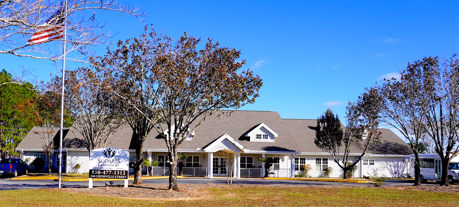 Exterior view of Sodalis Cantonment senior living facility building with a clear blue sky, several leafless trees in front, an American flag on a flagpole, and a sign displaying the facility name, phone number, and address.