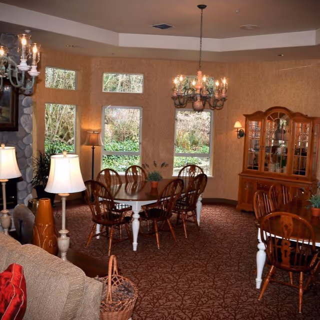 A cozy dining room with two round wooden tables surrounded by wooden chairs. The room has large windows showing greenery outside, a chandelier hanging from the ceiling, a wooden china cabinet, and several lamps providing warm lighting. The carpet has a patterned design, and there is a stone fireplace partially visible on the left.