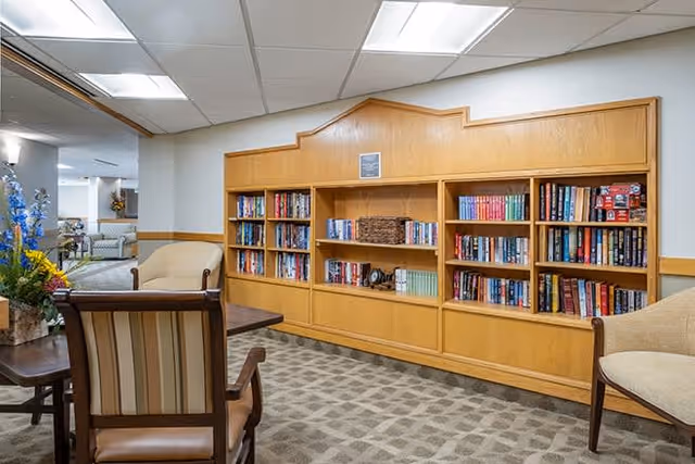A cozy reading area in a senior living facility featuring a built-in wooden bookshelf filled with books, two upholstered armchairs, a wooden table with a floral arrangement, and a carpeted floor. The space is well-lit with ceiling lights and has a neutral color scheme.