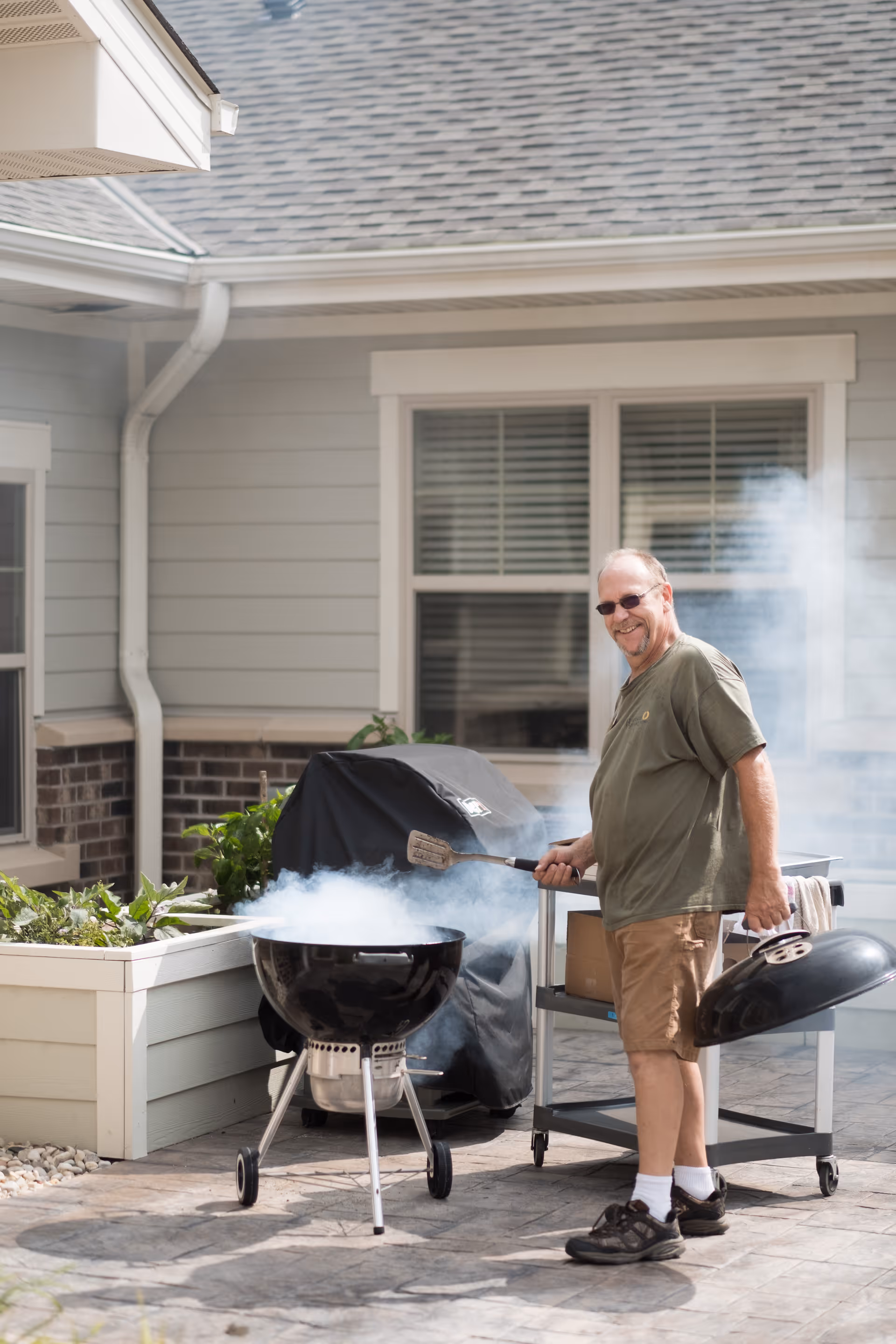 A man wearing sunglasses, a green t-shirt, and khaki shorts is standing outside on a patio next to a smoking charcoal grill. He is holding the grill lid in one hand and a spatula in the other, smiling at the camera. The background shows a light gray building with windows and some greenery in planter boxes.