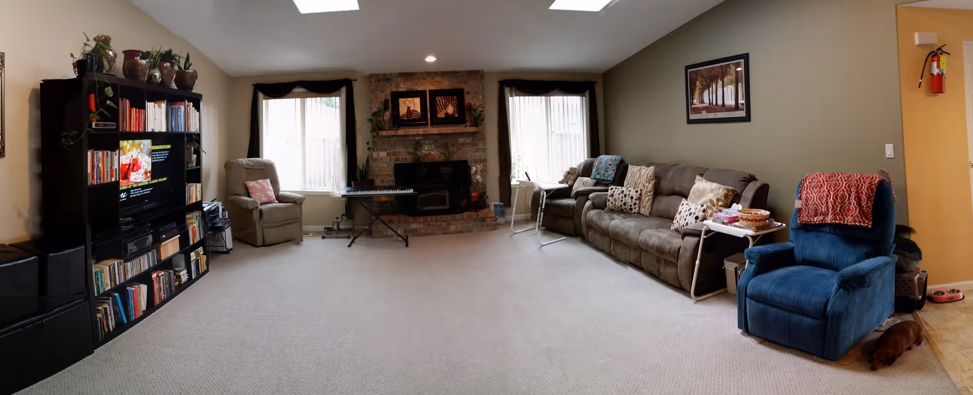 A spacious living room with beige carpet, featuring a large brown sofa with multiple patterned pillows, a blue recliner with a red throw blanket, and a beige armchair. There is a brick fireplace in the center with framed artwork above it, flanked by two windows with sheer curtains. A black entertainment center filled with books and a TV is on the left side. A small dog is lying on the floor near the blue recliner. The walls are painted in neutral tones, and there is a fire extinguisher mounted near the door on the right.