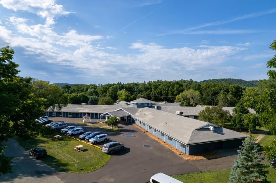 Aerial view of Eagles Rest Assisted Living facility showing a single-story building with a gray roof surrounded by trees and greenery. Several cars are parked in the parking lot adjacent to the building under a partly cloudy blue sky.
