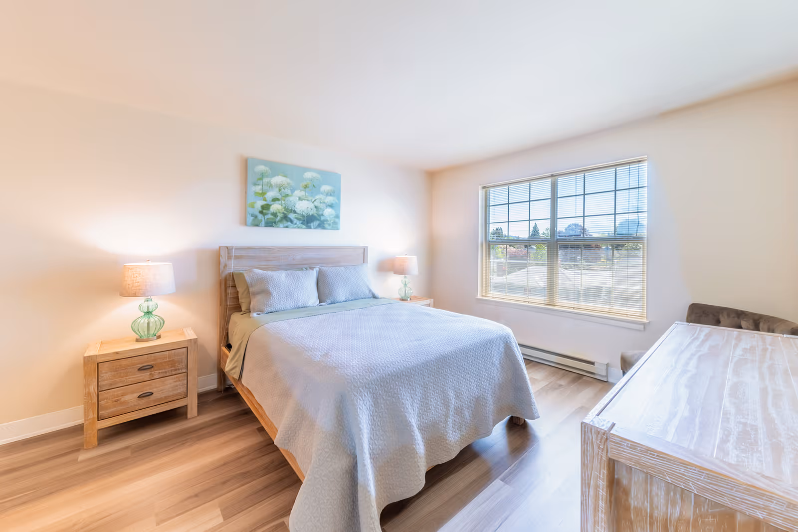A bright bedroom with a wooden bed frame, light gray bedding, two matching bedside tables with green glass lamps, a large window with blinds letting in natural light, a wooden dresser, and a floral painting on the wall above the bed.