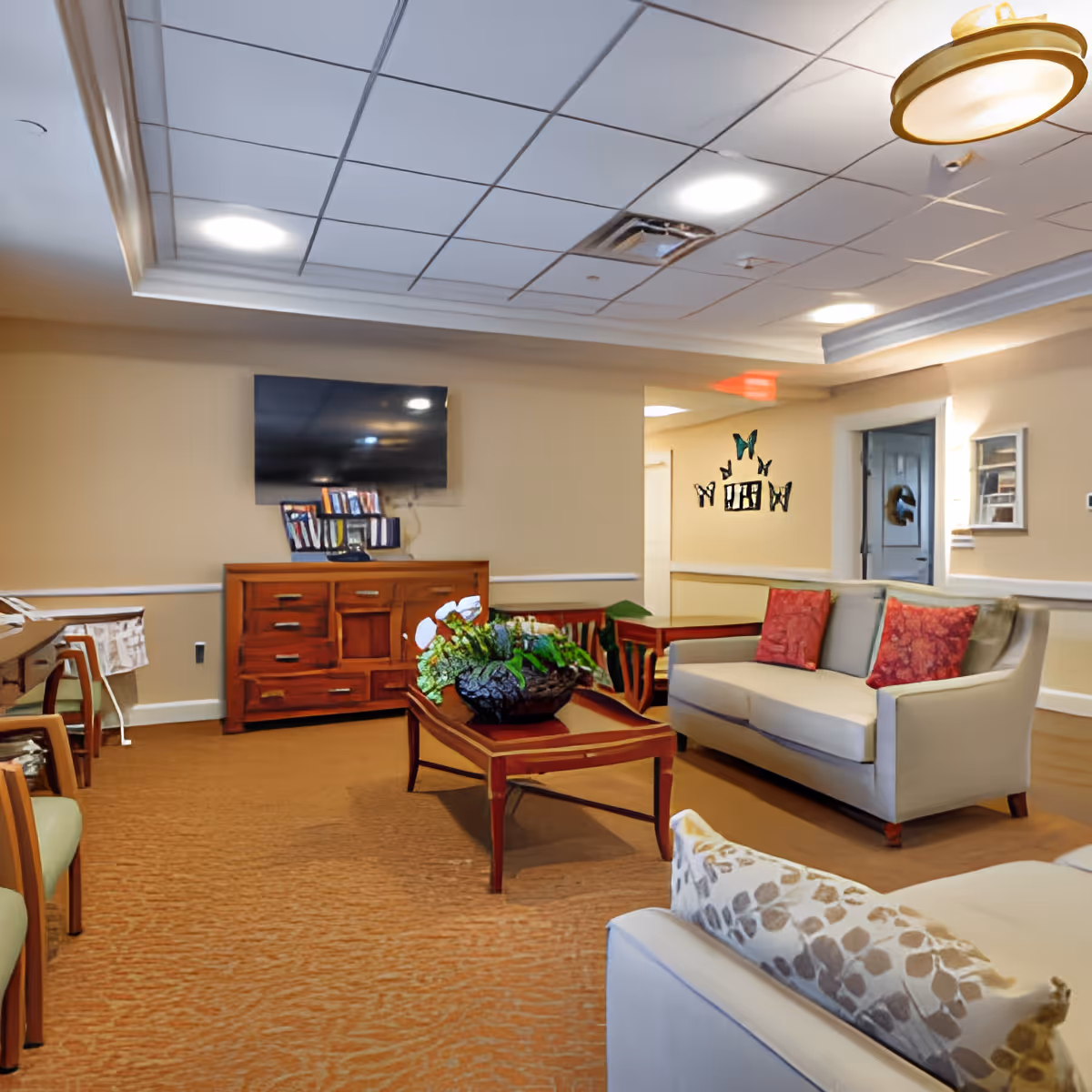 A cozy living room area in a senior living facility with beige walls and carpet. The room features a light gray sofa with red patterned pillows, a wooden coffee table with a decorative plant centerpiece, a wooden sideboard with books and a flat-screen TV mounted above it. There are additional chairs along the left side and butterfly wall decorations near a doorway.