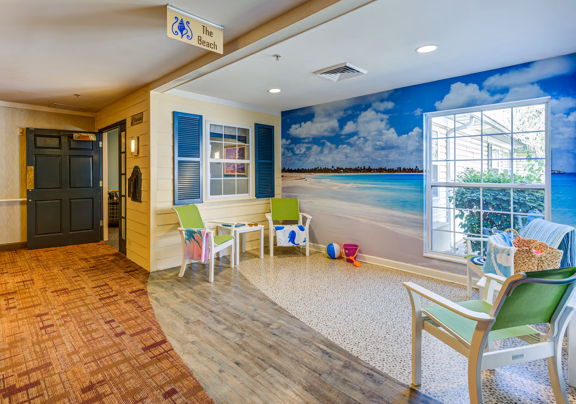 Indoor seating area designed to resemble a beach setting with a large wall mural of a sandy beach and ocean under a blue sky with clouds. The area includes white chairs with green cushions, a small white table, beach toys like a ball and bucket, and a window showing greenery outside. The floor transitions from wood to a pebble-like texture. A sign above reads 'The Beach.'