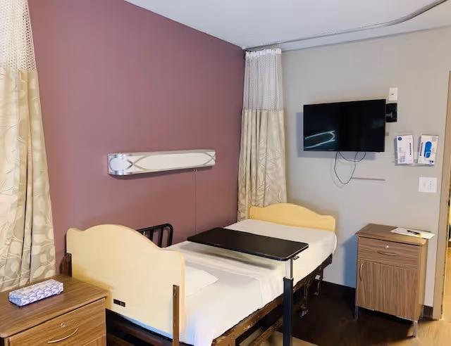 A small assisted-living patient room with a single adjustable bed, overbed table, wall-mounted TV, and bedside cabinets against a mauve accent wall.