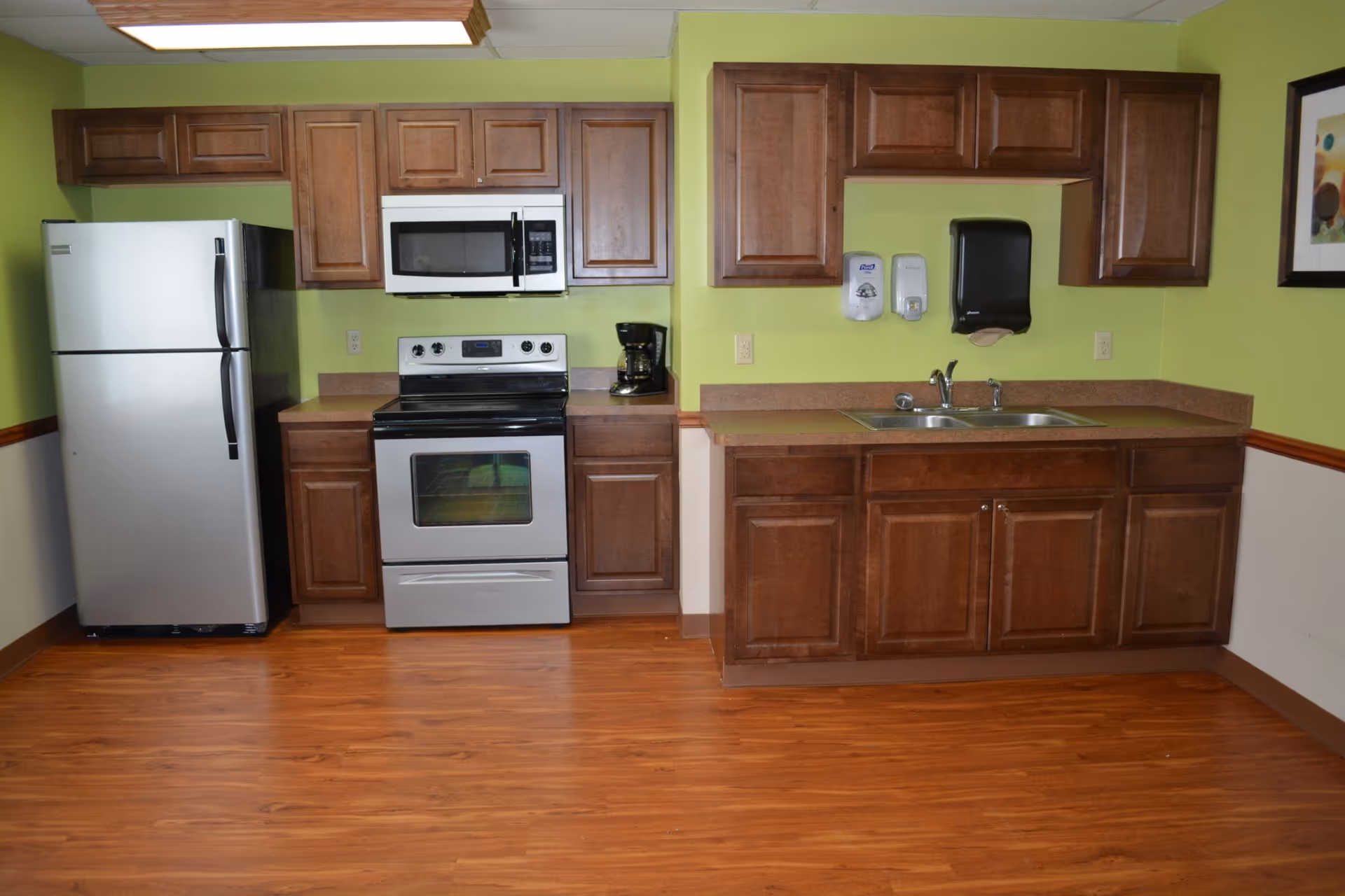 A kitchen area with wooden cabinets, a stainless steel refrigerator, an electric stove with an oven, a microwave above the stove, a coffee maker on the counter, and a double sink with soap dispensers and a paper towel dispenser mounted on the wall. The walls are painted light green and the floor is wood.