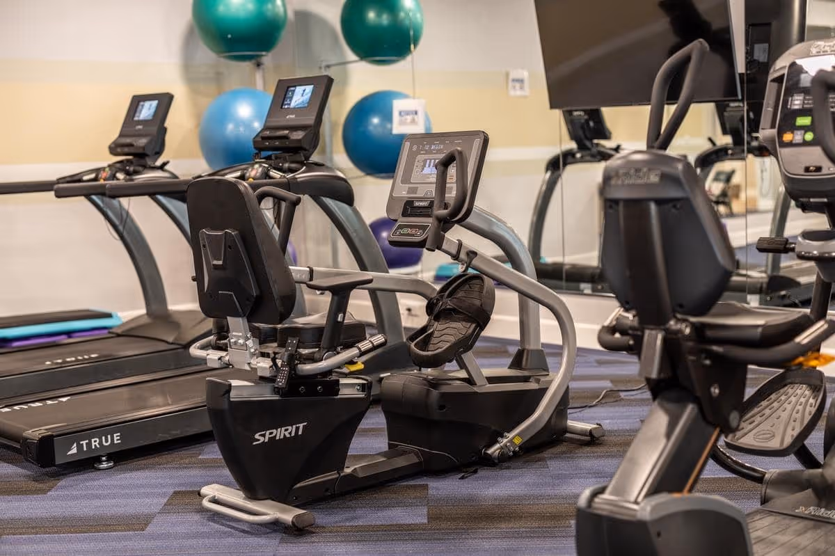 Indoor fitness room with exercise equipment including recumbent stationary bikes and treadmills, with exercise balls mounted on the wall in the background.