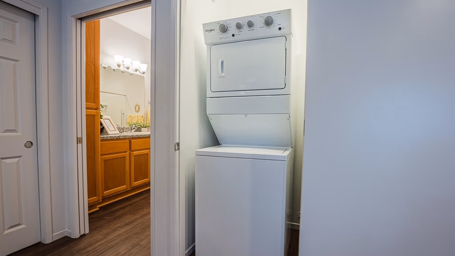 A stacked white washer and dryer unit in a small laundry closet with white walls and dark wood flooring. To the left, there is an open doorway leading to a bathroom with wooden cabinets, a granite countertop, a large mirror, and wall-mounted light fixtures.