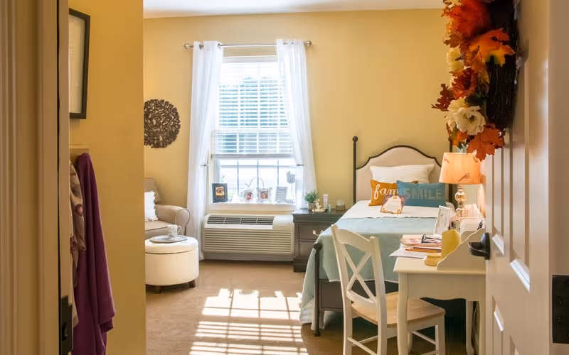 Sunlit furnished bedroom with a single bed, desk, armchair and a window with white curtains.