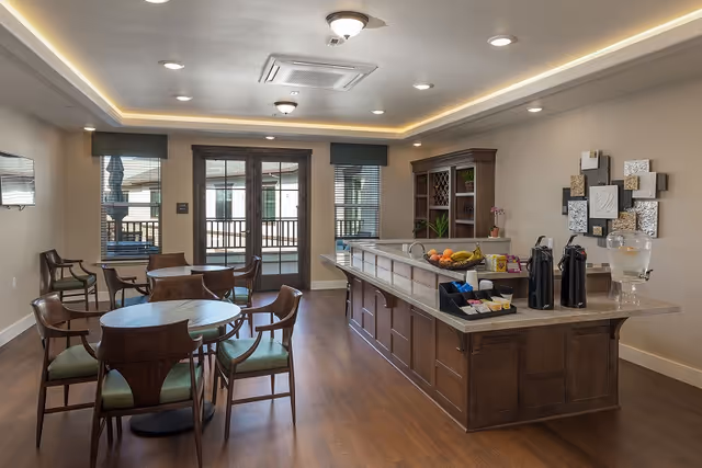 A well-lit dining area in a senior living facility featuring round wooden tables with green cushioned chairs, a long wooden counter with a granite top holding coffee dispensers, a water dispenser, fruit, and condiments. The room has large windows with blinds and a door leading to an outdoor balcony. The walls are decorated with a modern art piece and the floor is wooden.