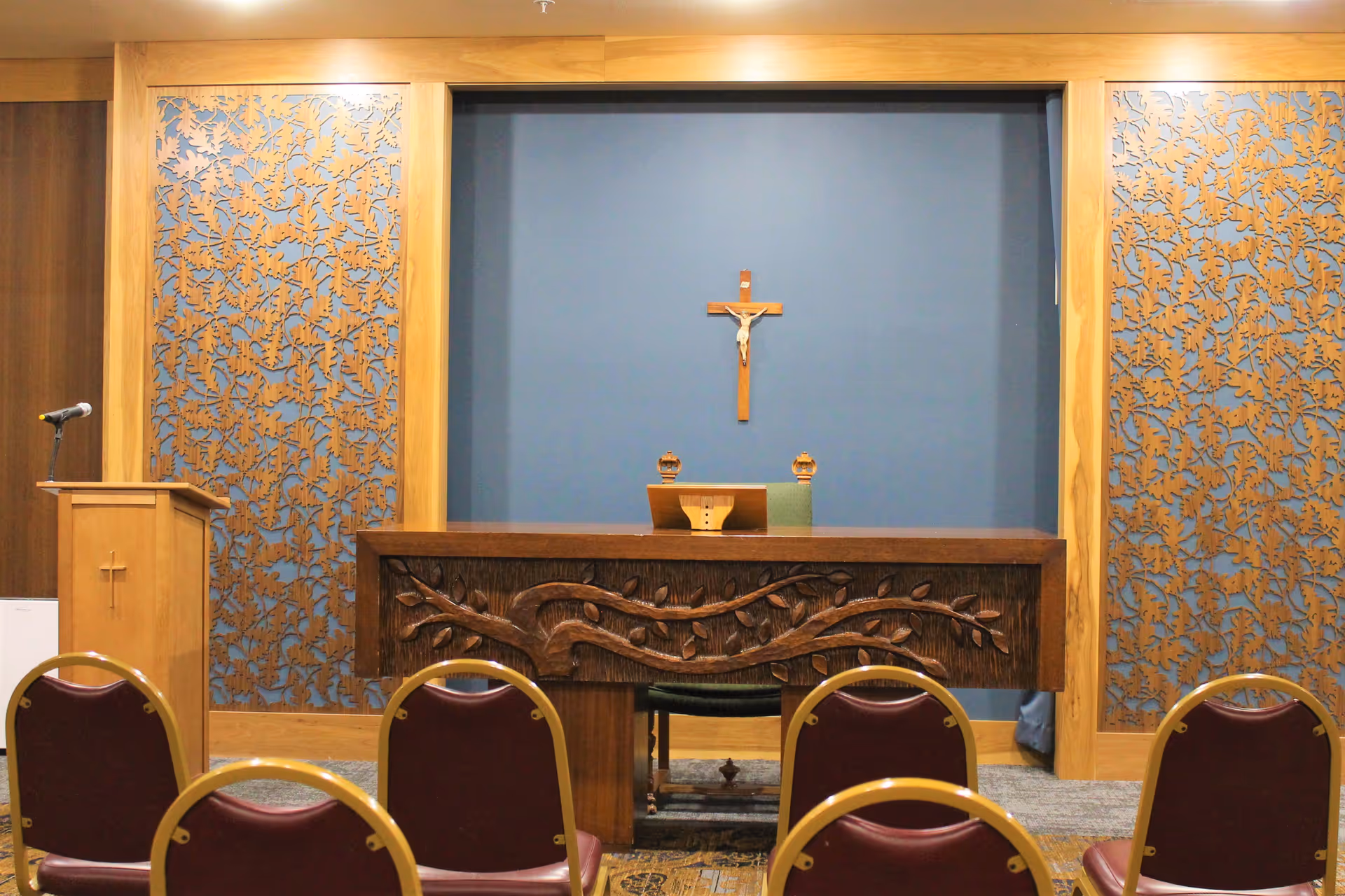 Small chapel interior with an altar carved with a vine motif, a crucifix on a blue wall, lectern, and rows of chairs.