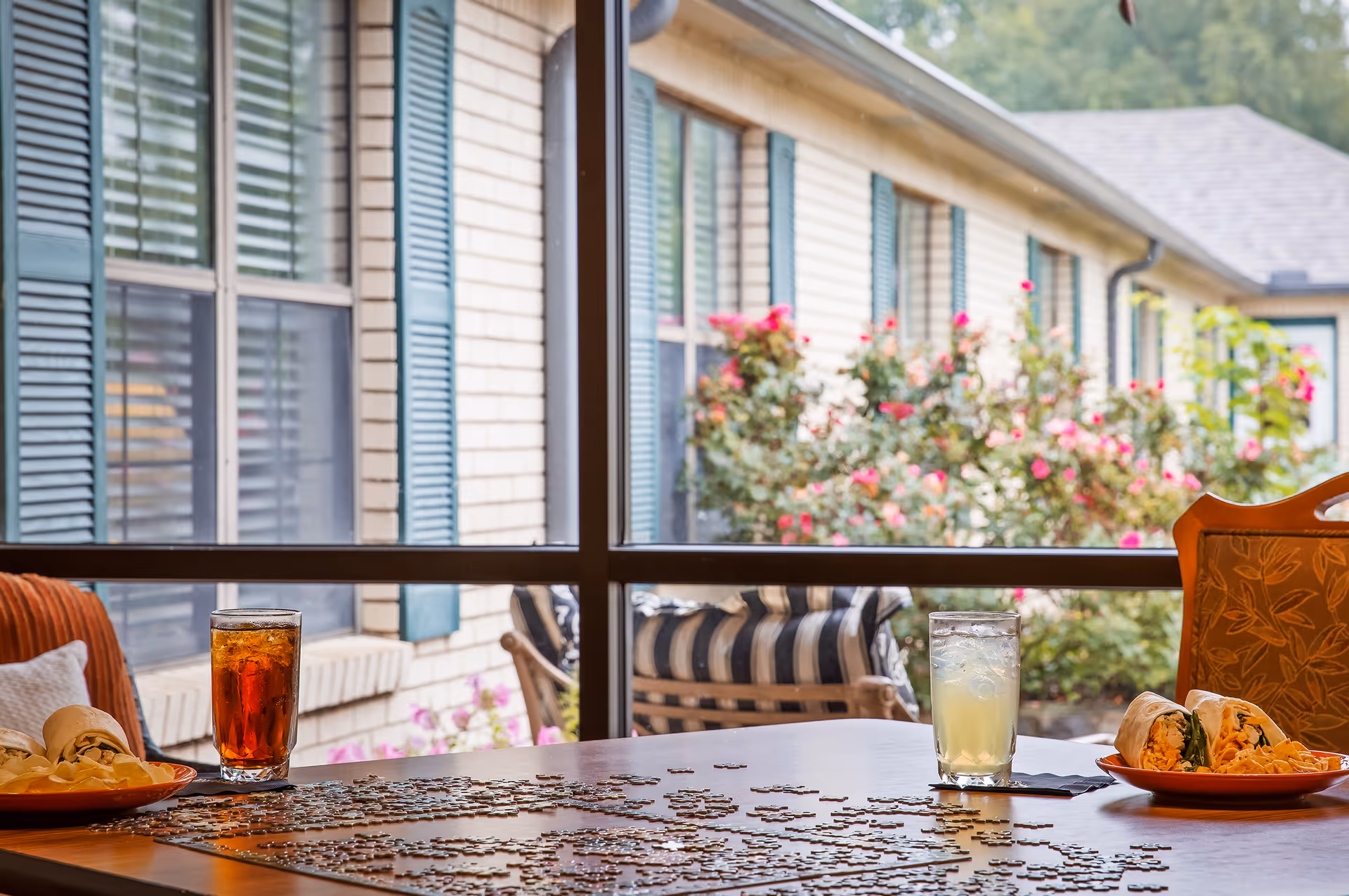 A dining table with drinks, plates of food and an unfinished jigsaw puzzle in front of a large window showing the building exterior and flowering bushes.