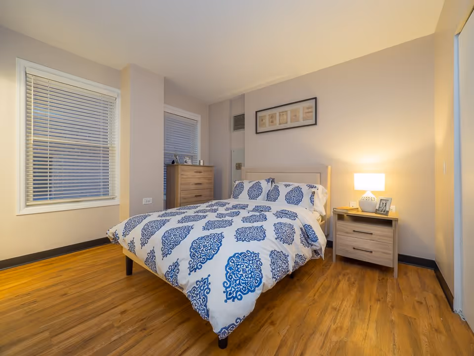 A cozy bedroom with a bed featuring white and blue patterned bedding. The room has wooden flooring, two windows with white blinds, a wooden dresser, and a wooden nightstand with a lit lamp and a framed photo. A framed artwork hangs above the bed.