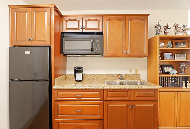 A kitchen area with wooden cabinets, a black refrigerator, a black microwave mounted above a granite countertop, a coffee maker on the counter, and a double sink. To the right, there is a wooden bookshelf with various decorative items and framed photos.