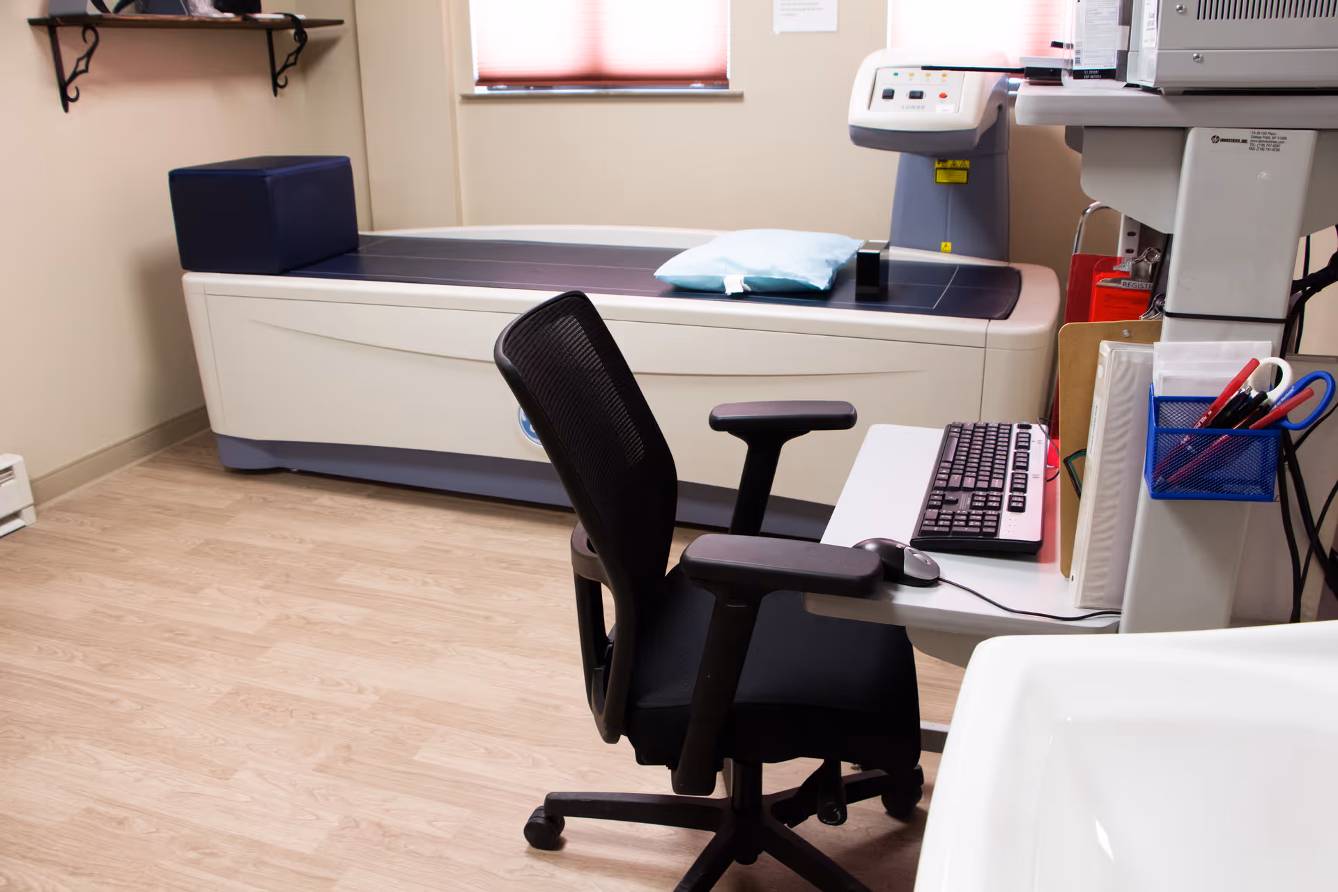 A medical examination room with a black office chair in front of a desk holding a keyboard and mouse. Behind the desk is a medical imaging machine with a padded examination table, a pillow, and a window with closed blinds in the background.