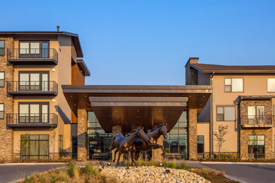 Exterior view of The Ridge Pinehurst senior living facility featuring a modern building with balconies, large windows, and a covered entrance. In front of the entrance, there is a sculpture of two horses on a rock bed surrounded by landscaping under a clear blue sky.