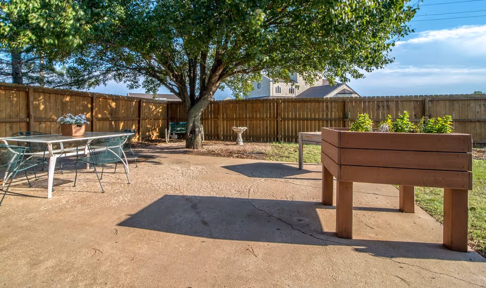 Outdoor patio area with a large tree providing shade, a wooden fence surrounding the space, a glass-top table with four metal chairs, a wooden raised garden bed with plants, a birdbath, and a bench in the background.