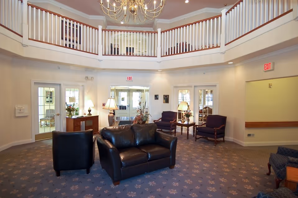 A spacious and well-lit common area in a senior living facility with a high ceiling and a chandelier. The room features a black leather loveseat and chair, two purple armchairs with wooden frames, side tables with lamps, and a carpeted floor with a floral pattern. There is a balcony railing on the upper level and multiple doors leading to other rooms.