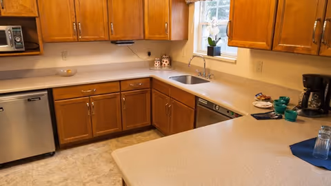A kitchen area with wooden cabinets, a stainless steel dishwasher, a microwave, a coffee maker, and a sink under a window. The countertops are light-colored and there are several teal cups and plates on the counter.