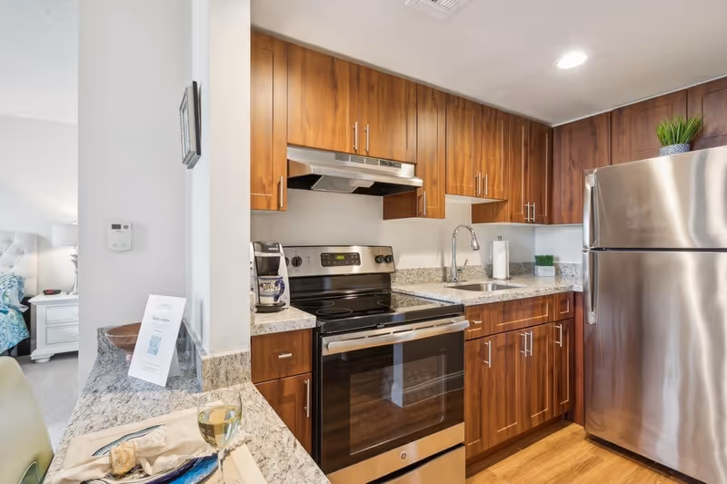 Modern kitchen with wooden cabinets, granite countertops, stainless steel refrigerator and electric stove with oven. A coffee maker is on the counter next to the stove. A small sink with a faucet is installed in the countertop. A glass of white wine and a plate with a napkin and fork are on the granite bar counter. Part of a bedroom with a white nightstand and lamp is visible through an open doorway.