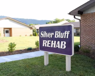 Outdoor view of a rehabilitation facility with a purple sign that reads 'Silver Bluff REHAB' on a grassy area near a sidewalk, with buildings and hills in the background.