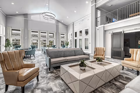 Spacious airy senior living lounge with high ceilings, large windows, sofas, armchairs and a central upholstered coffee table.