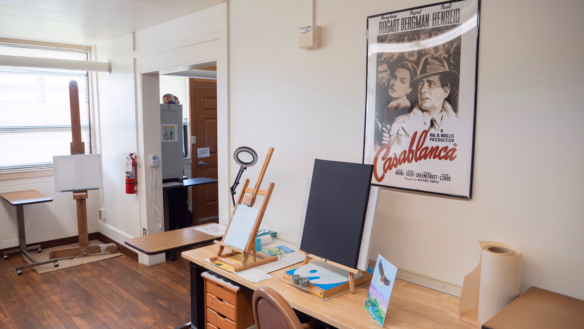 An art room with wooden floors and white walls featuring two easels with blank canvases, a desk with art supplies, a small painting of a bird, and a framed Casablanca movie poster on the wall. There is a window with blinds, a fire extinguisher, and a doorway leading to another room.