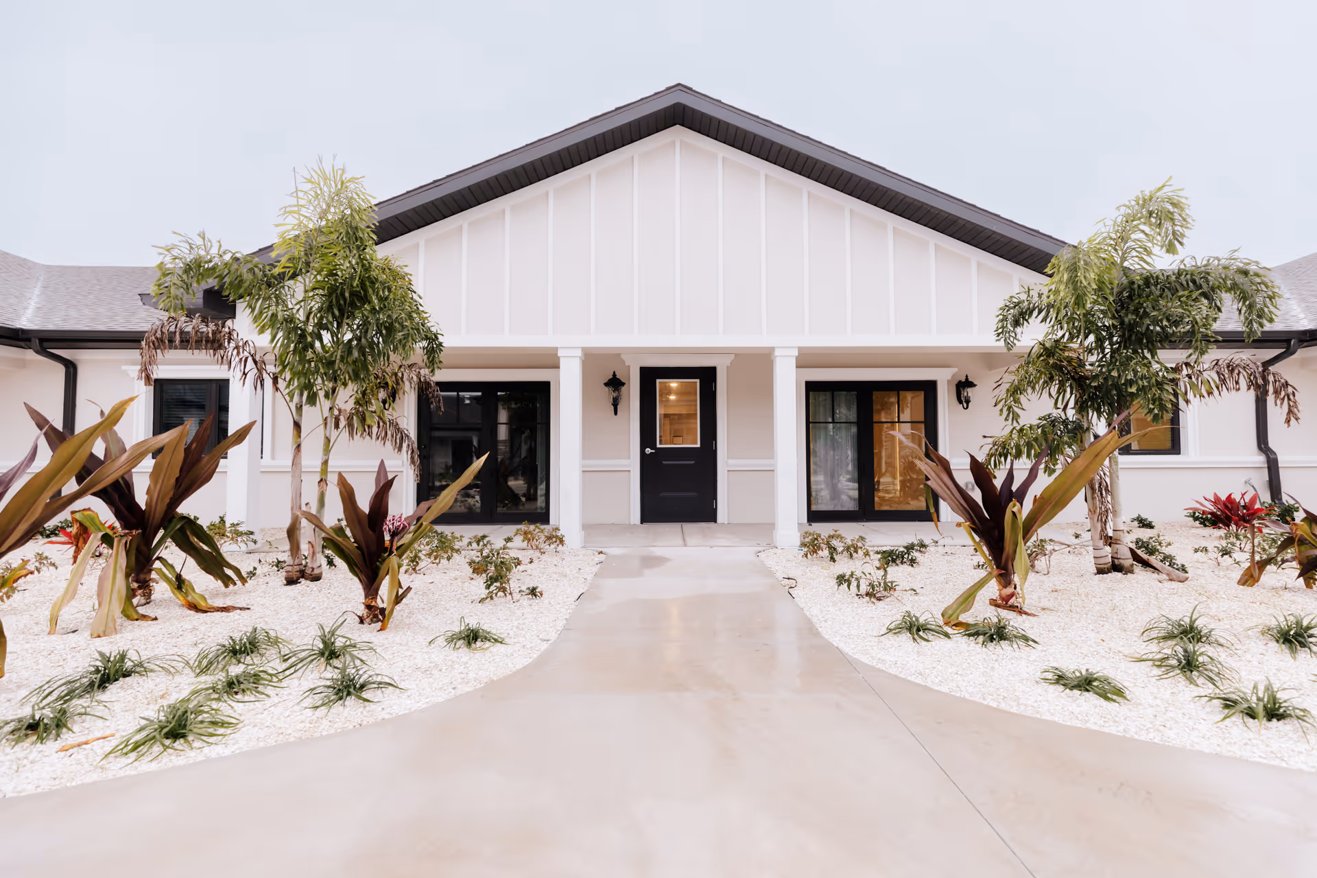Front exterior view of a single-story building with a white facade, black doors, and windows. The entrance is framed by two white columns and surrounded by landscaped garden beds with small palm trees and various plants on white gravel. The sky is overcast.