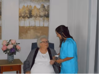 An elderly woman sitting in a chair holding hands with a caregiver dressed in blue scrubs in a room with a side table holding a vase of flowers and a painting of trees on the wall.