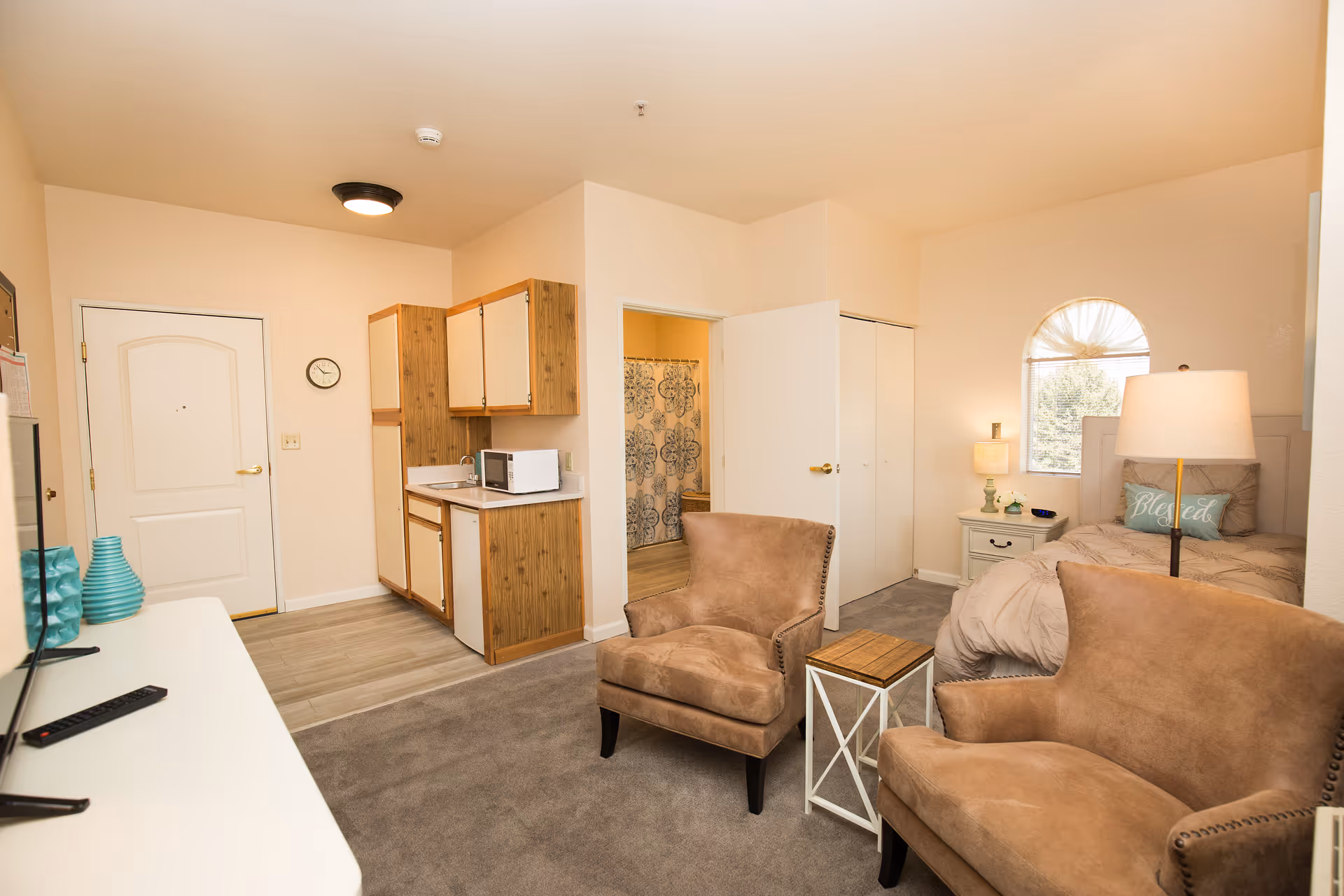 Interior view of a senior living facility room at Marycrest Assisted Living featuring a small kitchenette with wooden cabinets, a microwave, and a sink. The room includes two brown upholstered chairs with a small side table between them, a bed with a pillow that says 'Blessed,' a nightstand with a lamp, and a window with a decorative curtain. The entrance door and a bathroom with a shower curtain are visible in the background.