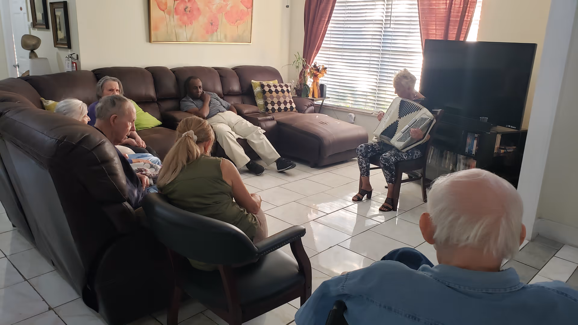 A group of elderly people seated in a living room area, attentively watching a woman playing an accordion near a window with blinds and red curtains. The room has a large brown sectional sofa, a TV on a stand, and tiled flooring.