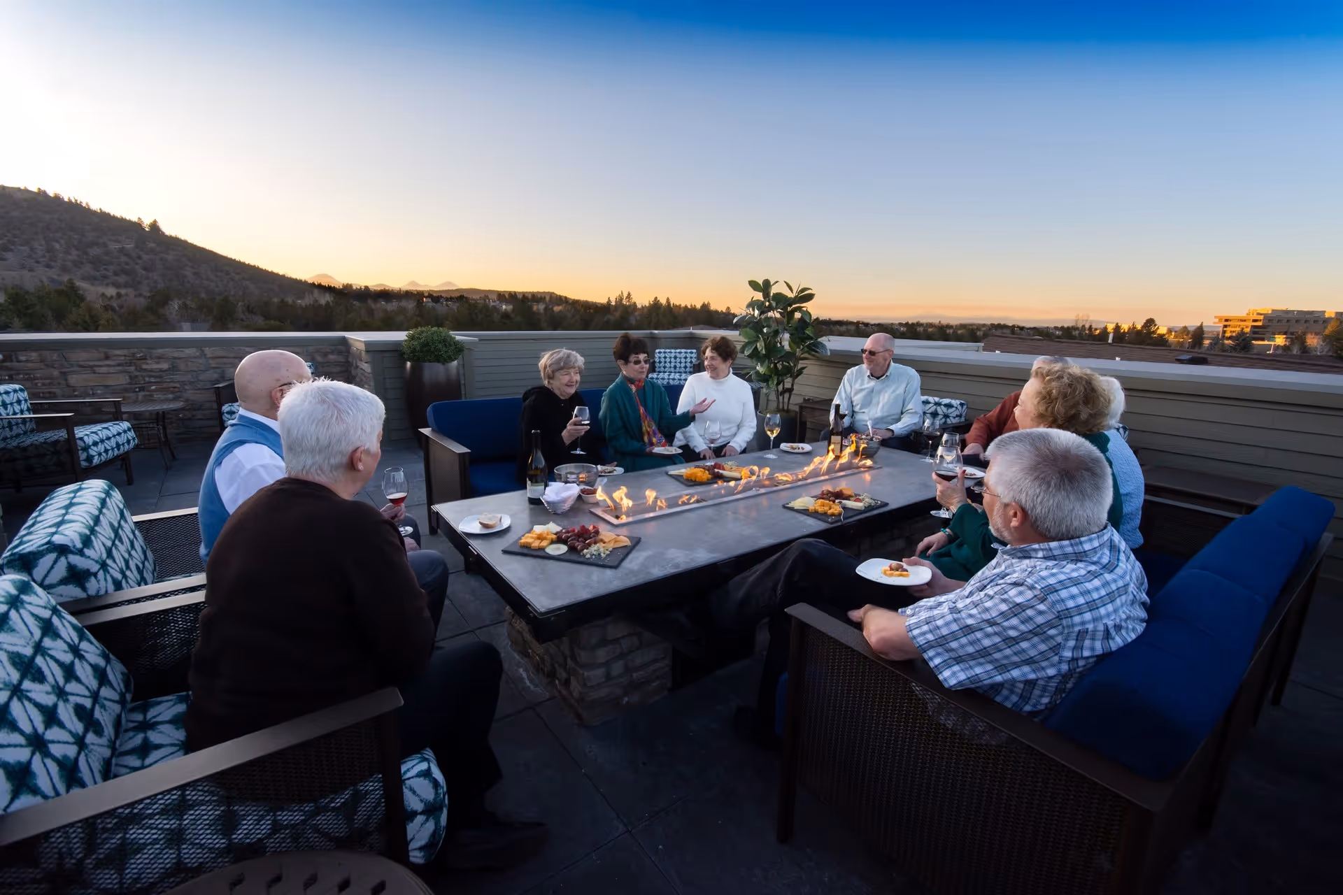 A group of elderly people sitting around a rectangular outdoor fire pit table on a rooftop patio during sunset, enjoying drinks and snacks while engaging in conversation. The seating includes cushioned chairs and sofas, with a scenic view of hills and trees in the background.