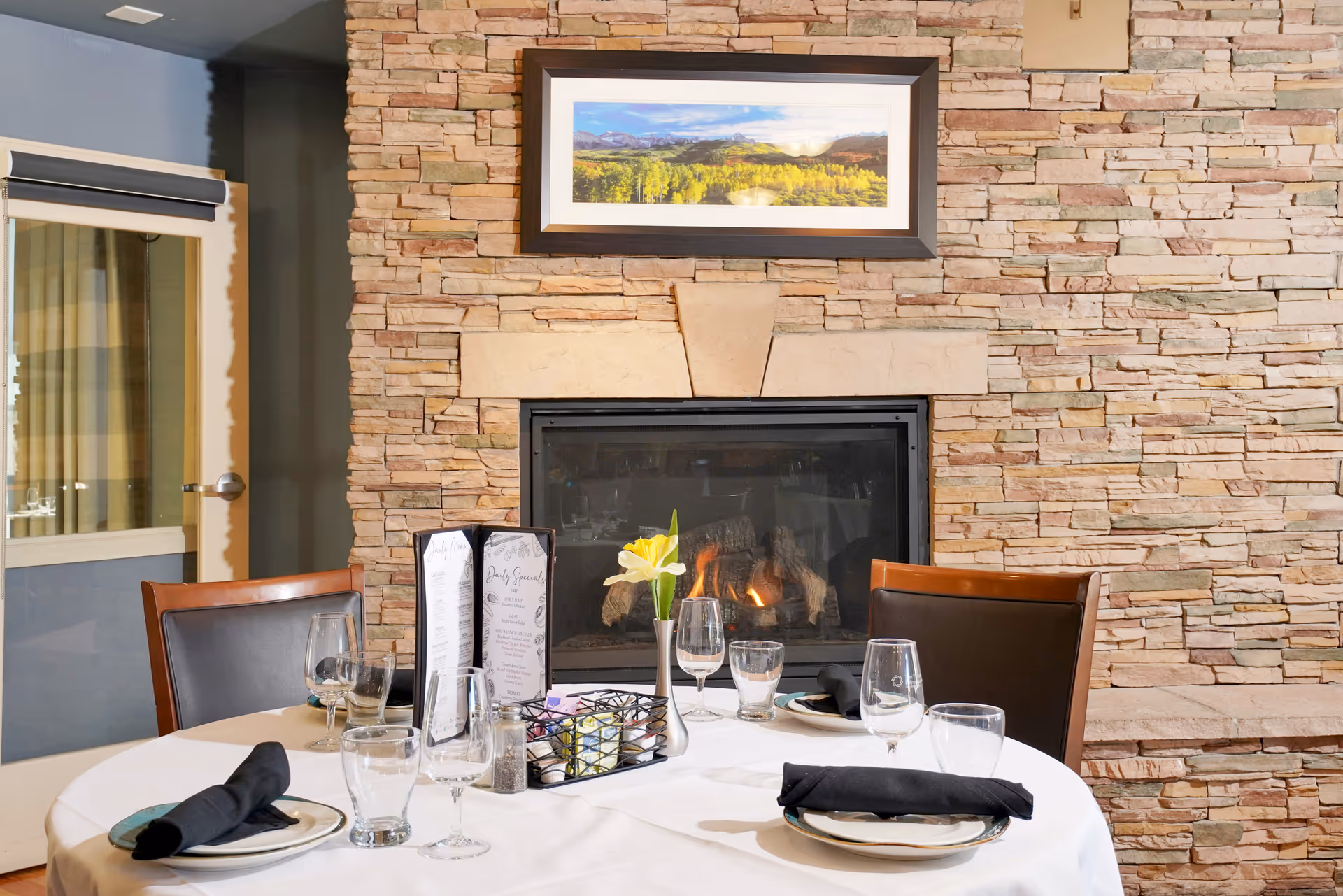 A dining table set for four with plates, glasses, and black napkins in front of a stone fireplace with a fire burning. A framed landscape picture hangs above the fireplace, and a door with a window is visible to the left.