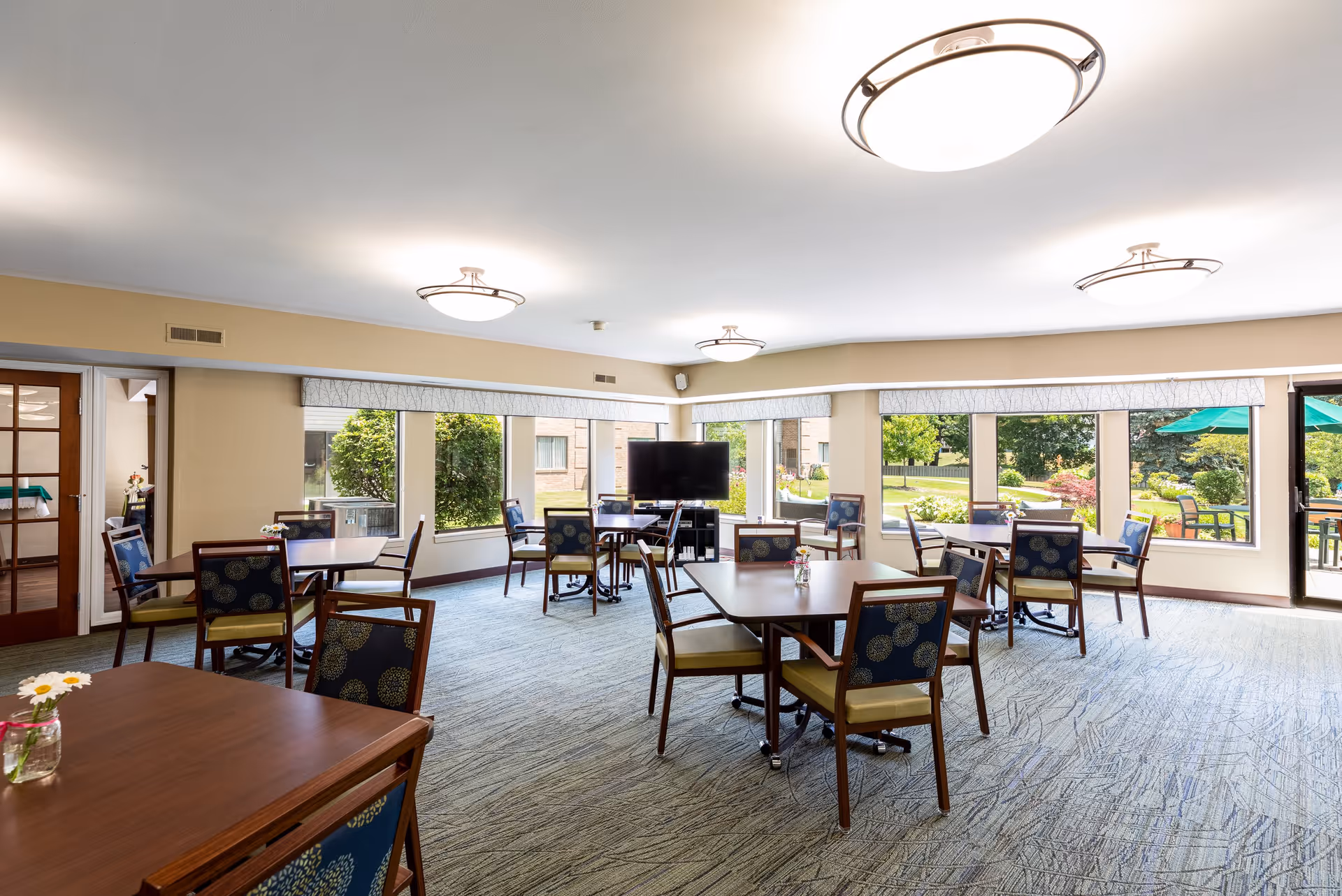 Interior view of a dining room with tables and chairs, large windows allowing natural light, and a television in the background. The room features a light color scheme and decorative elements.