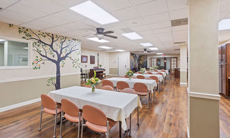 A dining room in a senior living facility with several tables covered in white tablecloths and decorated with flower vases. The room has wooden flooring, ceiling fans, and a mural of a tree painted on one wall. There are multiple chairs around the tables and a piano against the wall in the background.