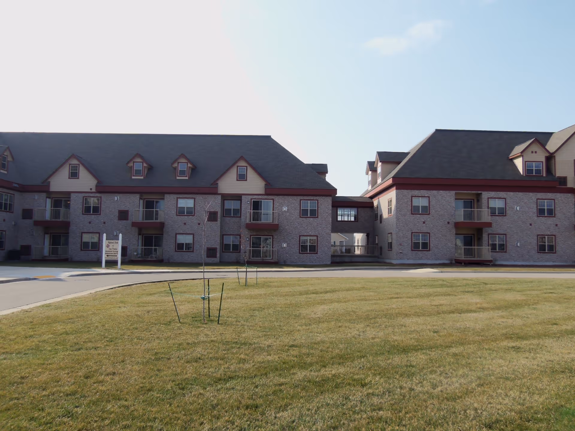 Exterior view of a two-story senior living facility building with brick walls, multiple windows, and small balconies. The building has a dark roof with dormer windows and is connected by a covered walkway. In front of the building is a well-maintained lawn with young trees and a paved driveway.