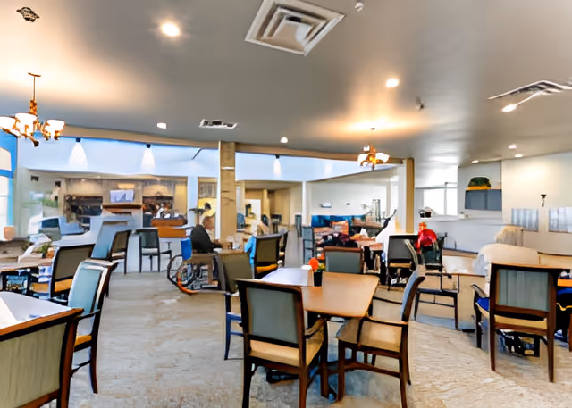 Communal dining and lounge area with multiple tables and chairs and several seated people, including a person in a wheelchair.