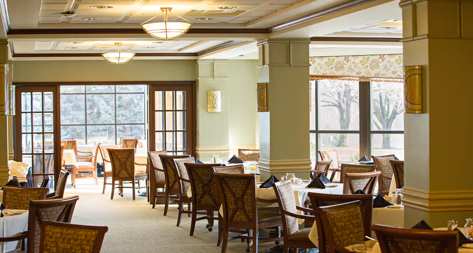 A bright dining room with multiple tables covered in white tablecloths and set with black napkins, surrounded by wooden chairs with patterned cushions. Large windows allow natural light to fill the room, and there are decorative columns and ceiling lights.
