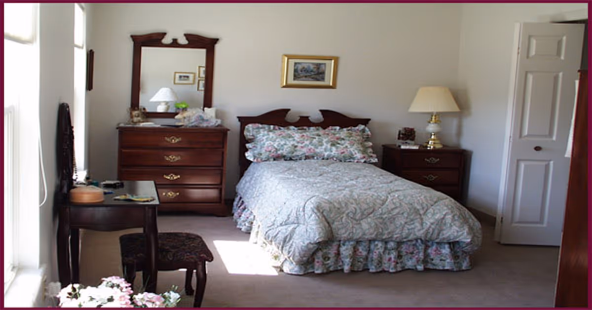 A cozy bedroom with a single bed covered in a floral bedspread and matching pillows. The room features a wooden dresser with a mirror, a nightstand with a lamp, a small table with a stool, and a partially open white door. There is a framed picture hanging on the wall above the bed and sunlight coming through a window on the left side.