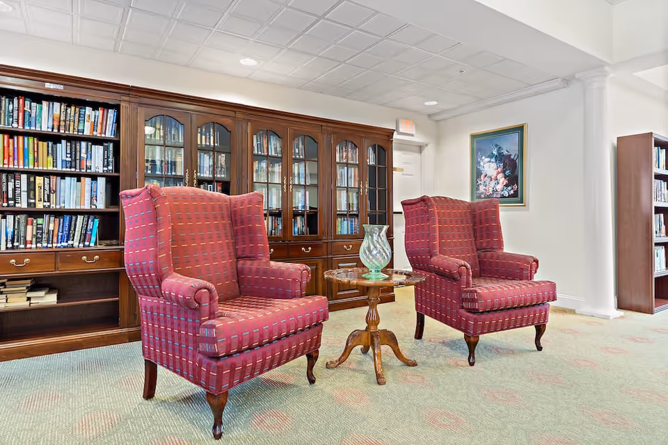 A cozy reading area in a senior living facility featuring two red patterned wingback chairs positioned around a small wooden table with a decorative glass vase. Behind the chairs is a large wooden bookshelf filled with books, and a framed floral painting hangs on the wall. The room has a light-colored carpet and a white ceiling with recessed lighting.